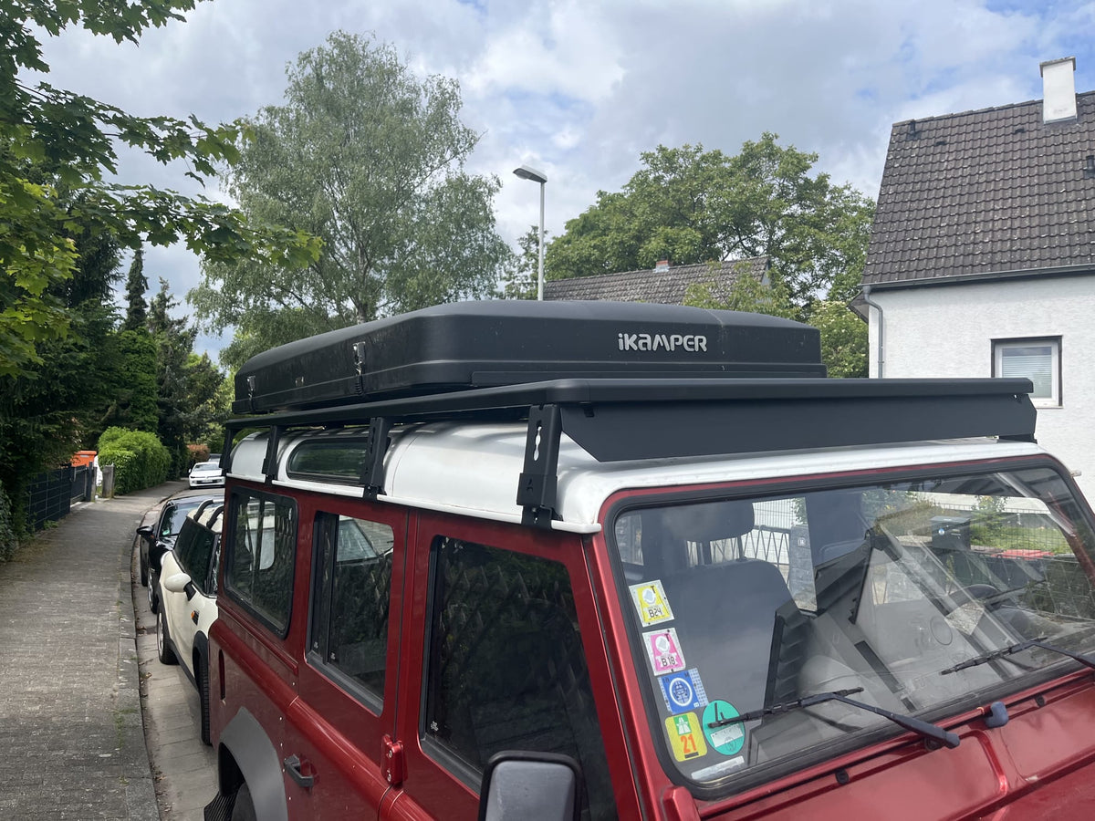Roter Land Rover Defender von vorne mit schwarzem modularen Airholder Dachträger inklusive Windabweiser und montiertem iKamper Hartschalen-Dachzelt.