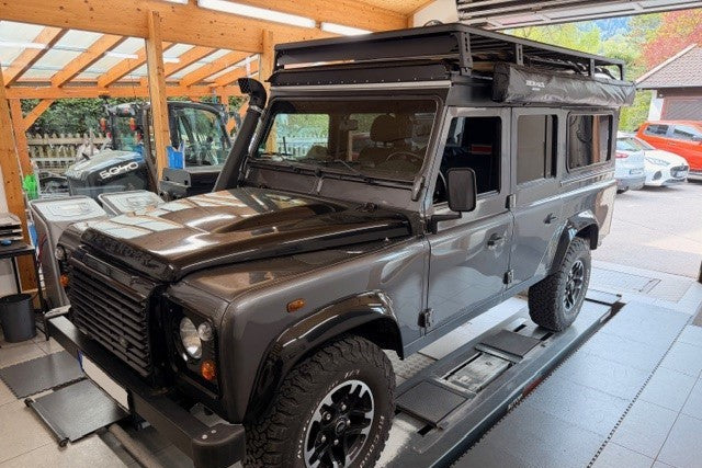 Grauer Land Rover Defender in einer Werkstatt mit montiertem Dachträger, Seitenmarkise und Ansaugschnorchel.