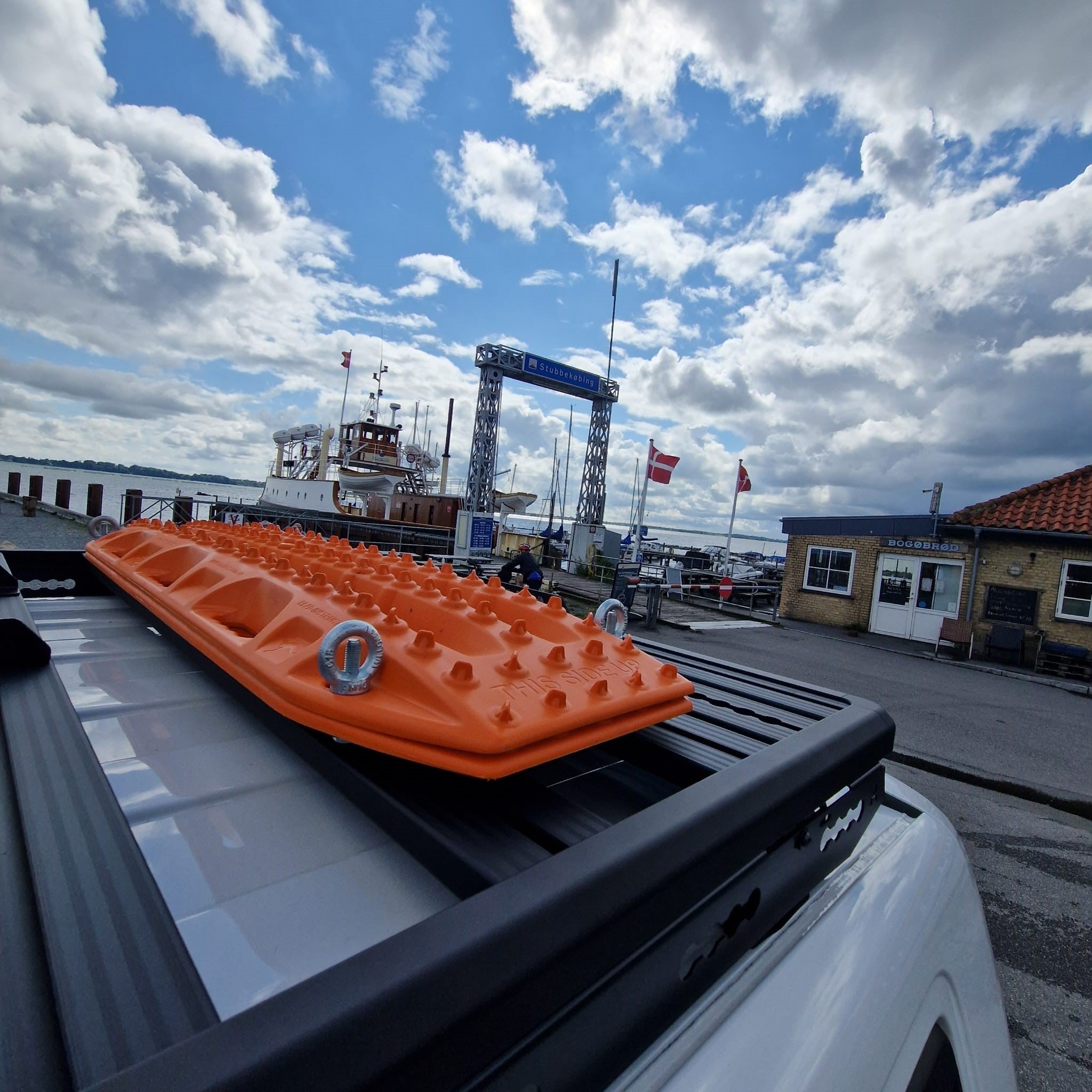 Orange Maxtrax-Bergeboards auf dem Airholder Dachträger eines weißen Fahrzeugs am Hafen montiert, im Hintergrund Boote und dänische Flaggen unter wolkigem Himmel.