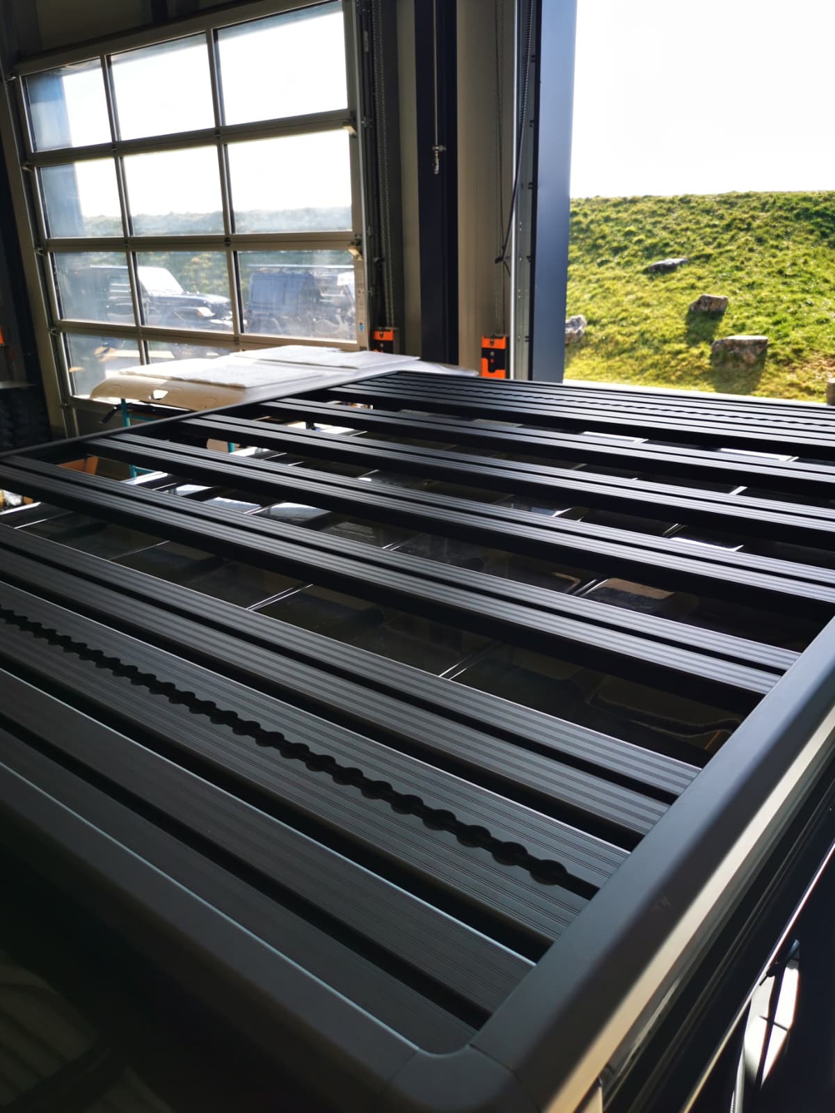 Airholder Dachträger-Plattform mit schwarzen Aluminium-Querprofilen in der Werkstatt, fotografiert am offenen Hallentor bei Tageslicht.