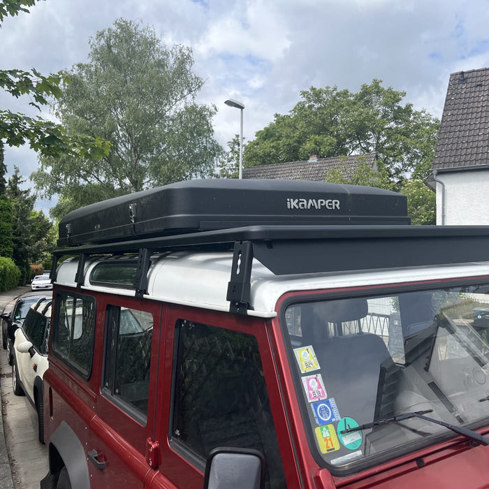 Roter Land Rover Defender von vorne mit schwarzem modularen Airholder Dachträger inklusive Windabweiser und montiertem iKamper Hartschalen-Dachzelt.