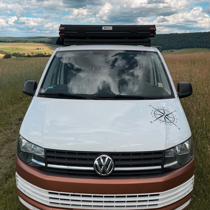 rontansicht eines weiß-braunen VW T6 mit einem flachen Airholder Dachträger und montiertem Hartschalen-Dachzelt, stehend auf einer Wiese vor einer Hügellandschaf