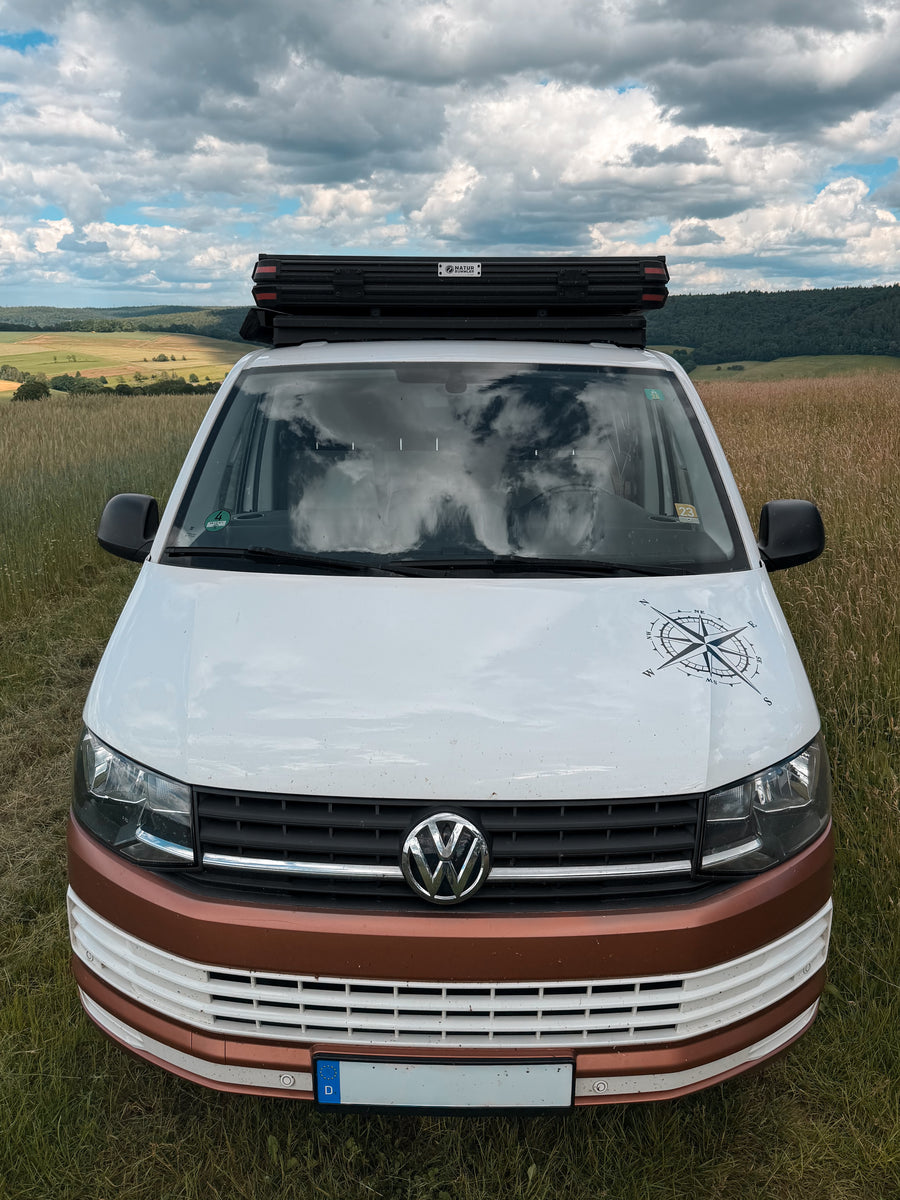rontansicht eines weiß-braunen VW T6 mit einem flachen Airholder Dachträger und montiertem Hartschalen-Dachzelt, stehend auf einer Wiese vor einer Hügellandschaf