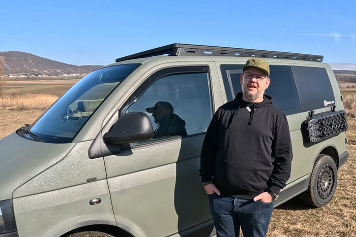 Ein olivgrüner VW Bus mit montiertem Dachträgersystem und Sandboards an der Seitenwand im Freien.